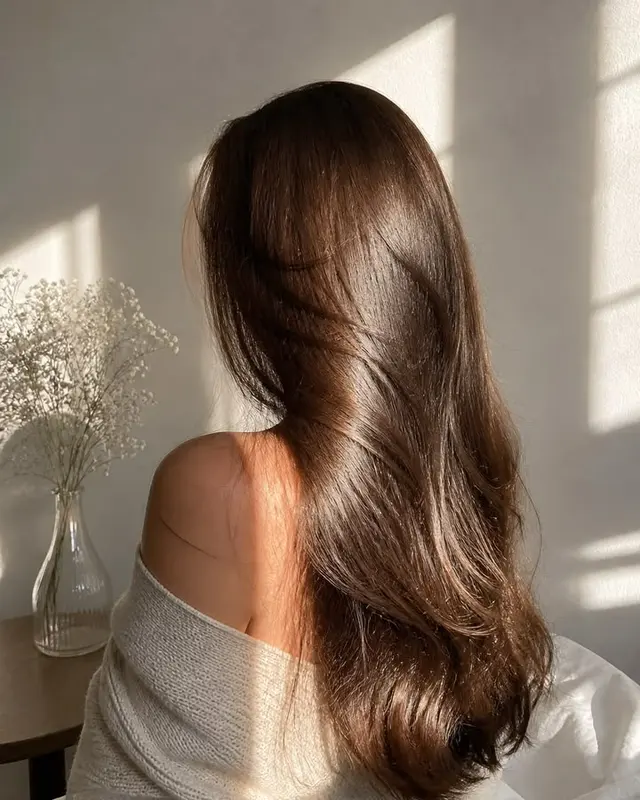 Mujer de espaldas con el cabello castaño largo, liso y brillante, junto a un florero con flores blancas