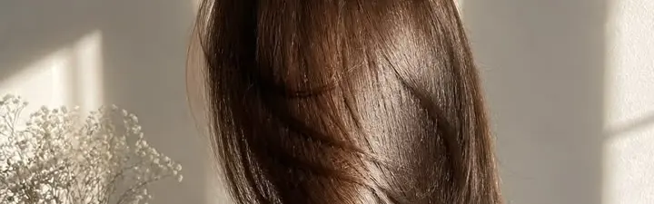 Mujer de espaldas con el cabello castaño largo, liso y brillante, junto a un florero con flores blancas