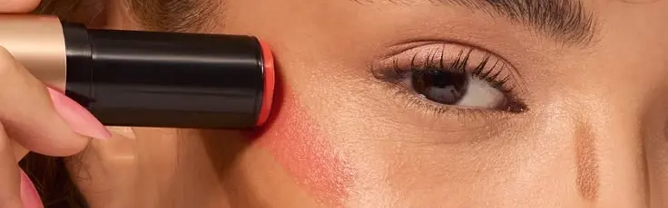 Mujer de cabello rizado aplicándose rubor en barra coral en la mejilla